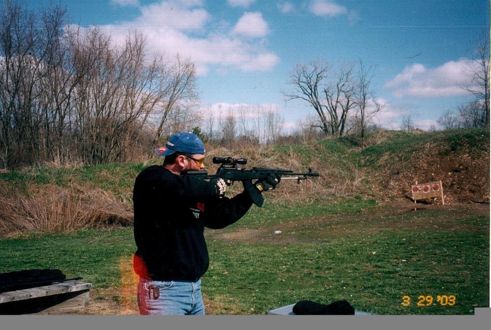 Man in black sweatshirt and blue cap aiming a scoped rifle at an outdoor shooting range