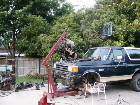 1991 Ford Bronco - A Ford Bronco is in a backyard with its engine being lifted out by an engine hoist, with parts scattered around.