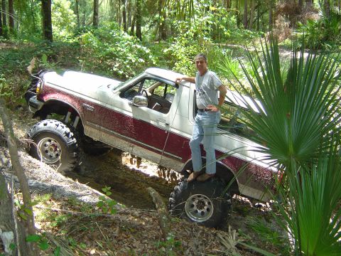 1993 Ford Bronco - A man in a gray T-shirt and jeans stands on the rear tire of a topless burgundy and white full-size Bronco tipped at a steep angle in a woo…