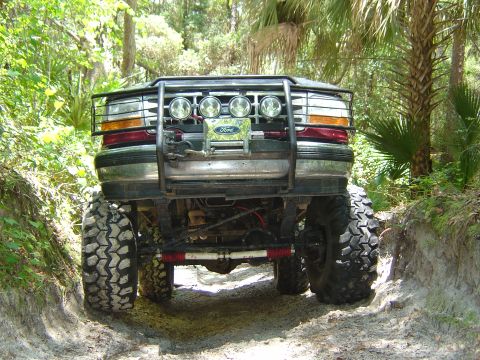 1993 Ford Bronco - Low front view of a lifted Ford 4x4 straddling a sandy rut, showing a black tube brush guard with four round driving lights, a Ford-badged…