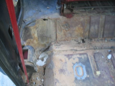 1981 Ford Bronco - The interior floor pan of a vehicle shows significant rust and damage, with a red seatbelt partially visible.