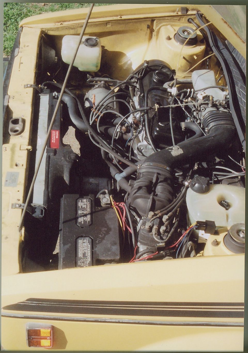Engine bay of a yellow 1979 VW Rabbit showing inline-4 engine, battery, hoses, and reservoirs.