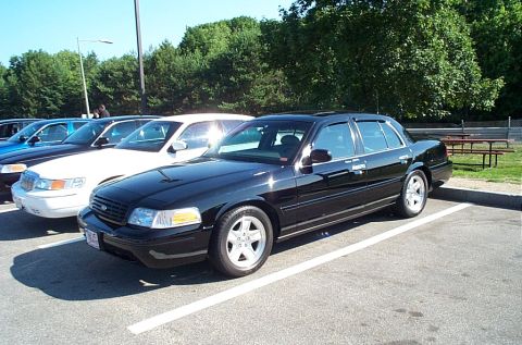 2001 Ford Crown Victoria - A black 2001 Ford Crown Victoria is parked in a lot, with several other vehicles visible in the background.