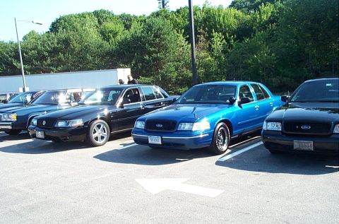 2001 Ford Crown Victoria - A bright blue Ford Crown Victoria on black steel wheels is parked between a black Mercury Marauder-style sedan with polished five-spoke whe…
