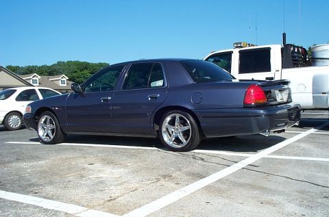 2001 Ford Crown Victoria - A dark gray/blue Ford Crown Victoria sedan viewed from the rear three-quarter angle, parked in a paved lot with aftermarket chrome five-spo…