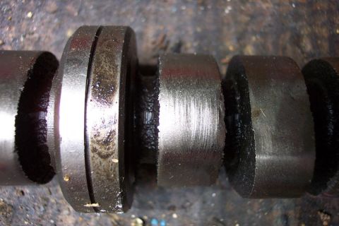 1968 Ford F-100 - Extreme close-up of a removed engine camshaft, showing several lobes and a bearing journal covered in dark oil residue. One lobe face shows…