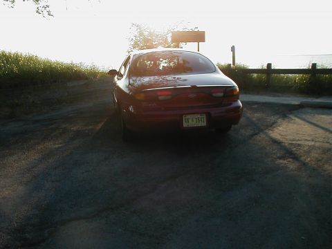 1997 Ford Taurus - Rear view of a red sedan parked near a wooden fence and grassy area at sunset.