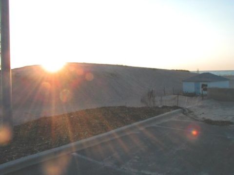 1997 Ford Taurus - The sun sets over a sandy dune, casting rays of light across the scene near a small beachside building.
