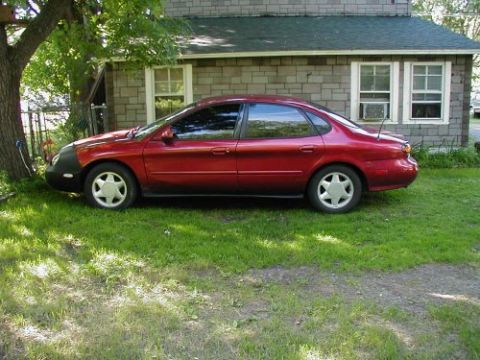 1997 Ford Taurus - A red sedan with a black fender cover sits on a grassy lawn next to a house.