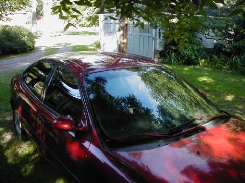 1997 Ford Taurus - A red sedan is parked in a grassy area next to a garage, partially shaded by trees.