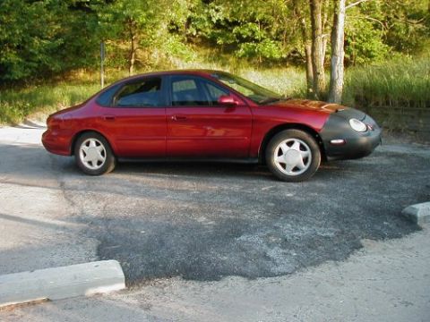 1997 Ford Taurus - A red sedan is parked in a lot with trees in the background. The front of the car is covered by a bra.