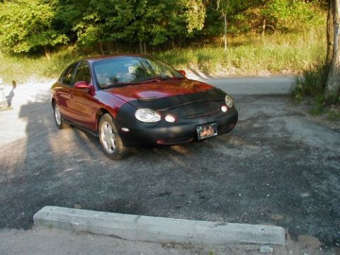 1997 Ford Taurus - A red 1997 Ford Taurus sedan is parked outdoors, partially covered by a black car bra.