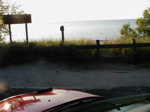 Red car hood in foreground, beach and water in background with a wooden fence and signs