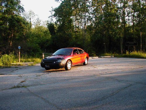 Red 1997 Ford Taurus sedan with a black car bra parked on asphalt, trees in background