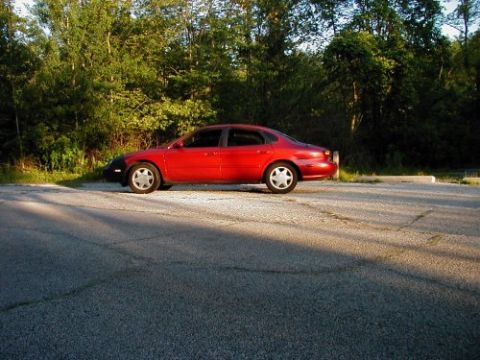 1997 Ford Taurus - A red 1997 Ford Taurus sedan is parked on a cracked asphalt lot with trees in the background.