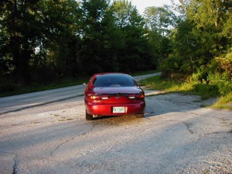 1997 Ford Taurus - A red 1997 Ford Taurus is seen from the rear on a gravel road with trees lining the sides.
