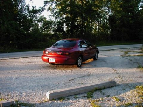 1997 Ford Taurus - Rear view of a red 1997 Ford Taurus sedan parked on a gravel lot with trees in the background.