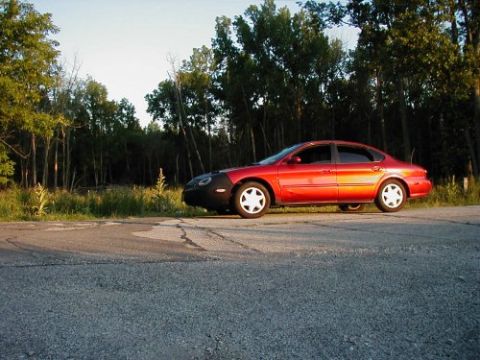 1997 Ford Taurus - A red sedan is parked on the side of a road with trees in the background.