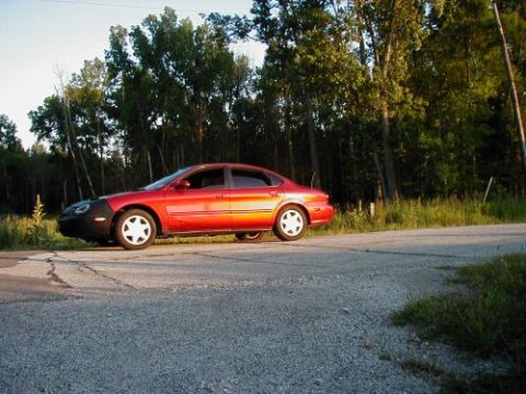 1997 Ford Taurus - A red 1997 Ford Taurus sedan is parked on the side of a road with trees in the background.
