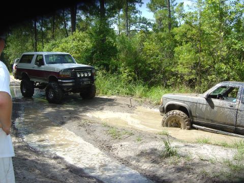 1993 Ford Bronco - Two modified trucks navigate a muddy off-road path, with one vehicle partially submerged in the mire.