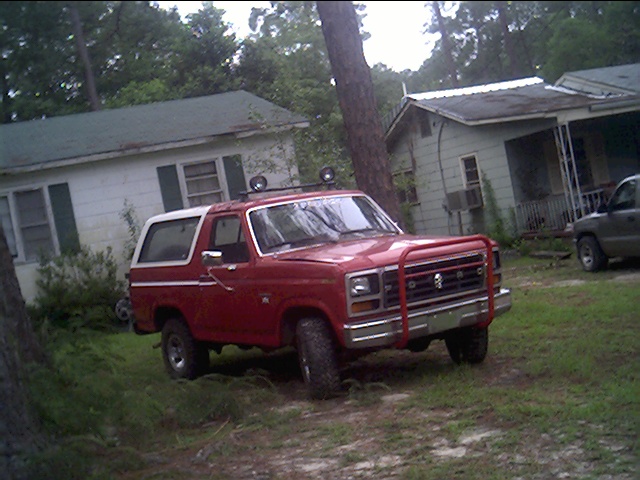 Red Ford Bronco with white top, bull bar, and roof lights parked on grass near houses