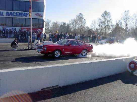 1976 Ford Mustang II doing a burnout on a drag strip, with a crowd watching
