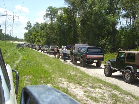 1993 Ford Bronco - A convoy of off-road vehicles, including a 1993 Ford Bronco, travels down a dirt path lined with trees and power lines.