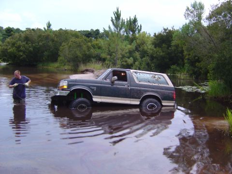 1993 Ford Bronco - Man attempts to free a 1993 Ford Bronco stuck in a body of water surrounded by trees.