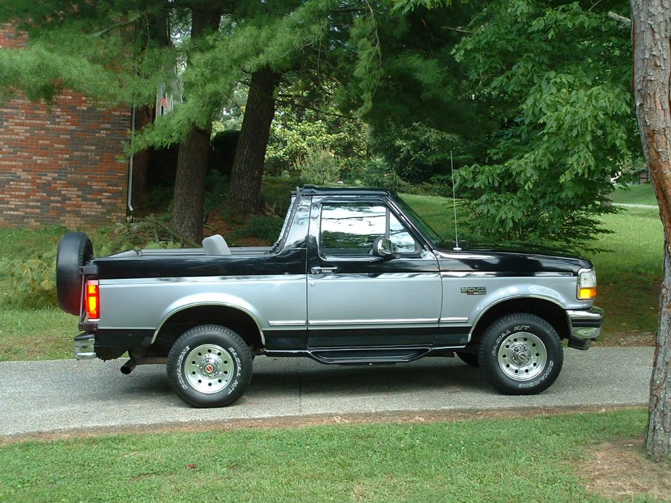 Black and silver Ford Bronco with hardtop removed, side view, parked on a driveway among trees