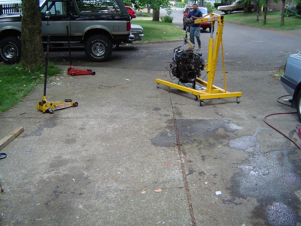 Engine hanging from a yellow gantry crane in a driveway, floor jacks and a Bronco parked nearby