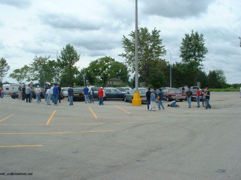 2000 Ford Crown Victoria - Wide shot of a car-club gathering in a large empty parking lot under overcast skies, with people standing among a row of parked full-size s…
