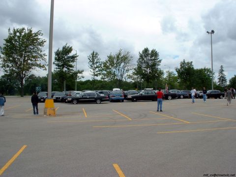 2000 Ford Crown Victoria - A club cruise-in staged in a wide, mostly empty asphalt parking lot under grey clouds, with a line of dark full-size sedans including Ford…