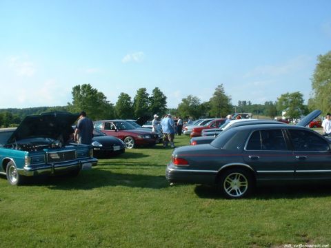 2000 Ford Crown Victoria - Outdoor car show on a grass field with a grey Mercury Grand Marquis on gold-face alloys and a red pinstripe in the foreground right, a teal…