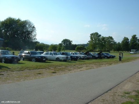 2000 Ford Crown Victoria - A row of Ford Panther-platform sedans parked nose-out on grass at an outdoor car meet, including black Crown Victorias and Grand Marquis an…