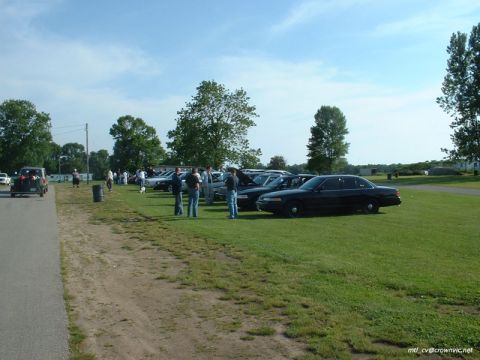 2000 Ford Crown Victoria - A black Ford Crown Victoria parked on grass in the foreground of a sunny club meet, with more sedans lined up beyond it and small groups of…
