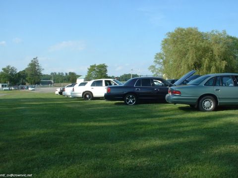 2000 Ford Crown Victoria - Low side view along a row of Panther-platform sedans parked on a mown grass field, with a silver-green Ford Crown Victoria on lacy alloys i…