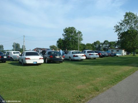 2000 Ford Crown Victoria - Rear view down a long row of full-size sedans parked on grass at a sunny club gathering, led by a white sedan, a black Ford Crown Victoria…