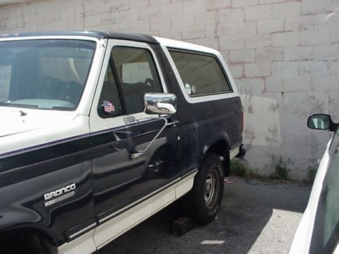 1989 Ford Bronco - A two-tone Ford Bronco is parked next to a brick wall, with another vehicle partially visible on the right.