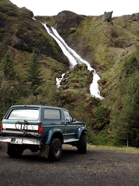 1996 Ford Bronco - A 1996 Ford Bronco is parked on a gravel road with a scenic waterfall cascading down a mossy hillside in the background.