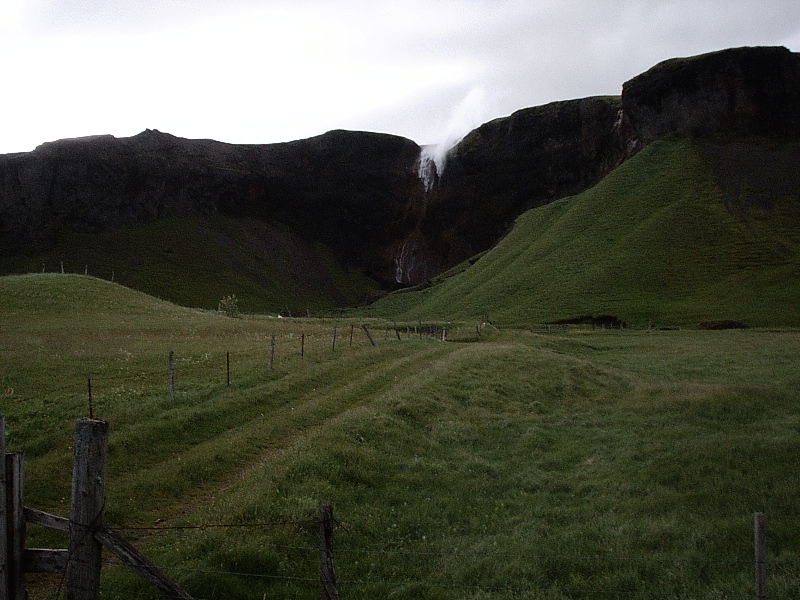 Green valley with dirt track leading to cliffs and a waterfall under gray sky