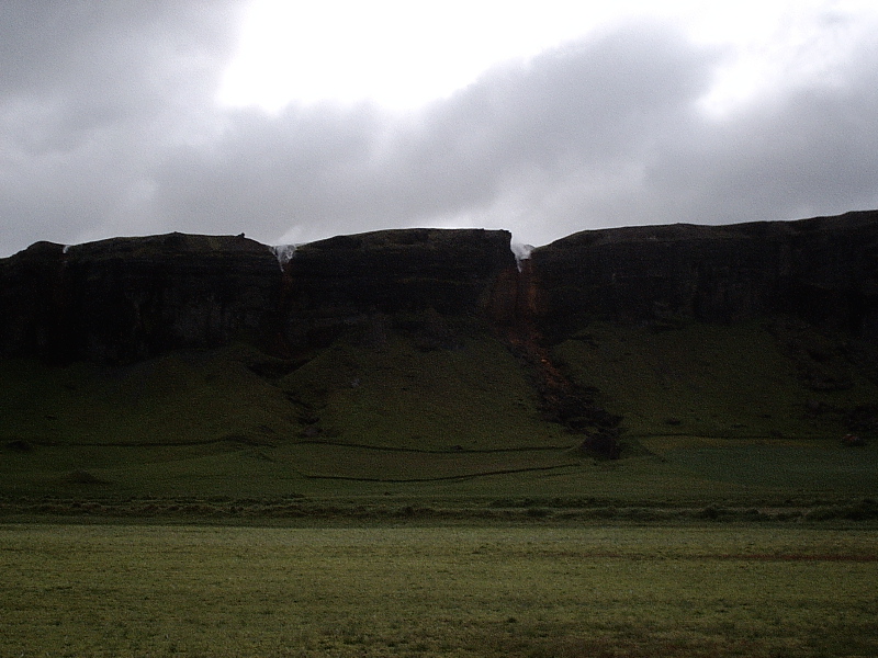 Green cliff with thin waterfalls under a gray overcast sky.