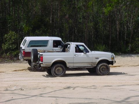 1986 Ford Bronco - Two white Ford Broncos are parked on a dirt road with trees in the background.