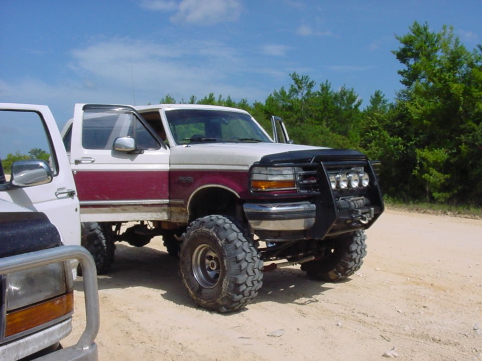 Lifted 1992 Ford Bronco with maroon stripe and bull bar, parked next to another truck