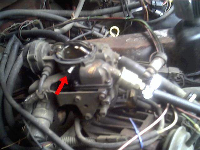 Close-up of a vehicle's engine carburetor with a red arrow pointing to a part