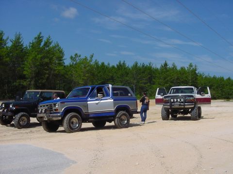 1986 Ford Bronco - Three off-road vehicles, including a blue and silver SUV, a Jeep, and a pickup truck, are parked on a dirt lot under a bright blue sky.