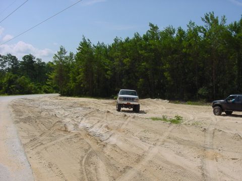 1986 Ford Bronco - Two trucks are parked on a sandy dirt road surrounded by trees under a clear blue sky.