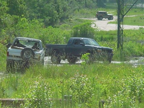 1986 Ford Bronco - Several vehicles navigate a muddy terrain surrounded by lush greenery, with one vehicle in the distance.