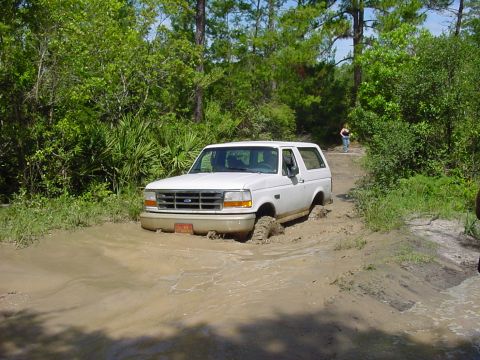 1986 Ford Bronco - A white Ford Bronco navigates a muddy trail, with its front tires deep in the mire.