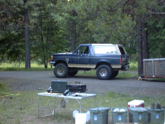 Lifted blue Ford Bronco parked at a wooded campsite with folding table and gear