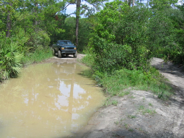 Dark green lifted Ford Bronco on a sandy trail behind a large muddy water hole